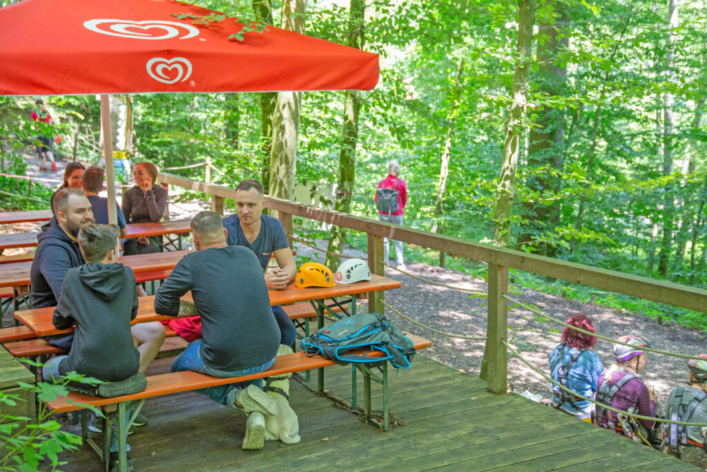 Biertische mit Personen, welche eine Pause vom Klettern machen. Die Personen reden miteinander. Die Tische sind im Kletterwald Prien aufgebaut und mit roten Sonnenschirmen überdacht.