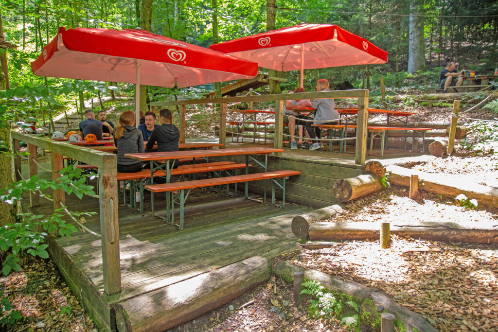 Zwei kleine Terrassen im Kletterwald Prien am Chiemsee. Auf dieser stehen Bierbänke sowie Biertische, auf denen Personen sitzen und sich ausruhen. Es sind zwei rote Sonnenschirme aufgespannt. Auf den Tischen liegen die Kletterhelme.