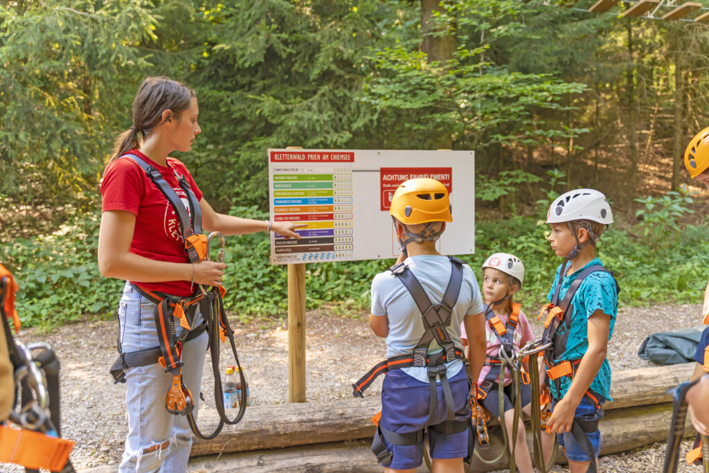Kinder in Kletterausrüstung beobachten eine Frau, währrend sie ihnen die Parcours an einer Tafel im Kletterwald zeigt.