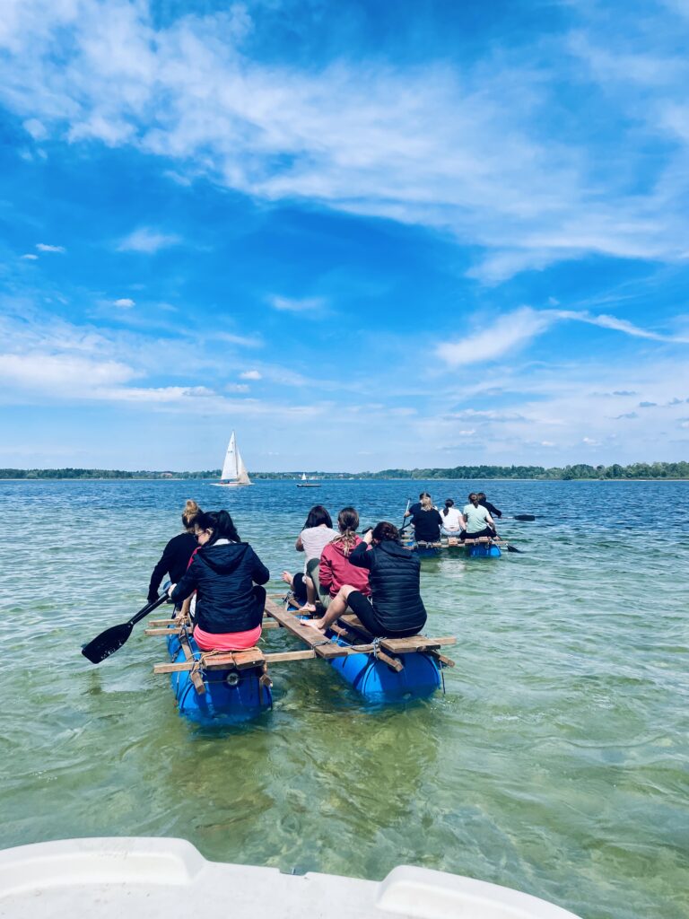 Sechs Personen sitzen auf einem selbstgebauent Floß, mit dem sie über den Chiemsee fahren.