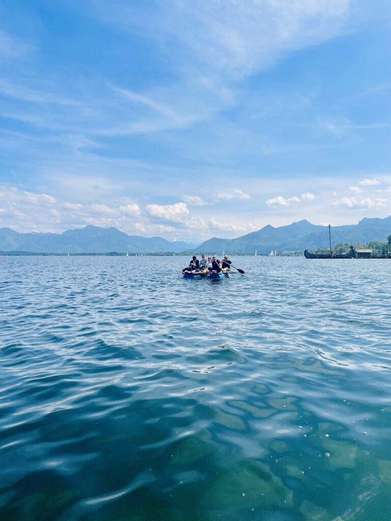 Fünf Personen sitzen auf einem selbstgebauten Floß und fahren damit an einem sonnigen Tag auf dem Chiemsee. Im Hintergrund sind die Alpen.
