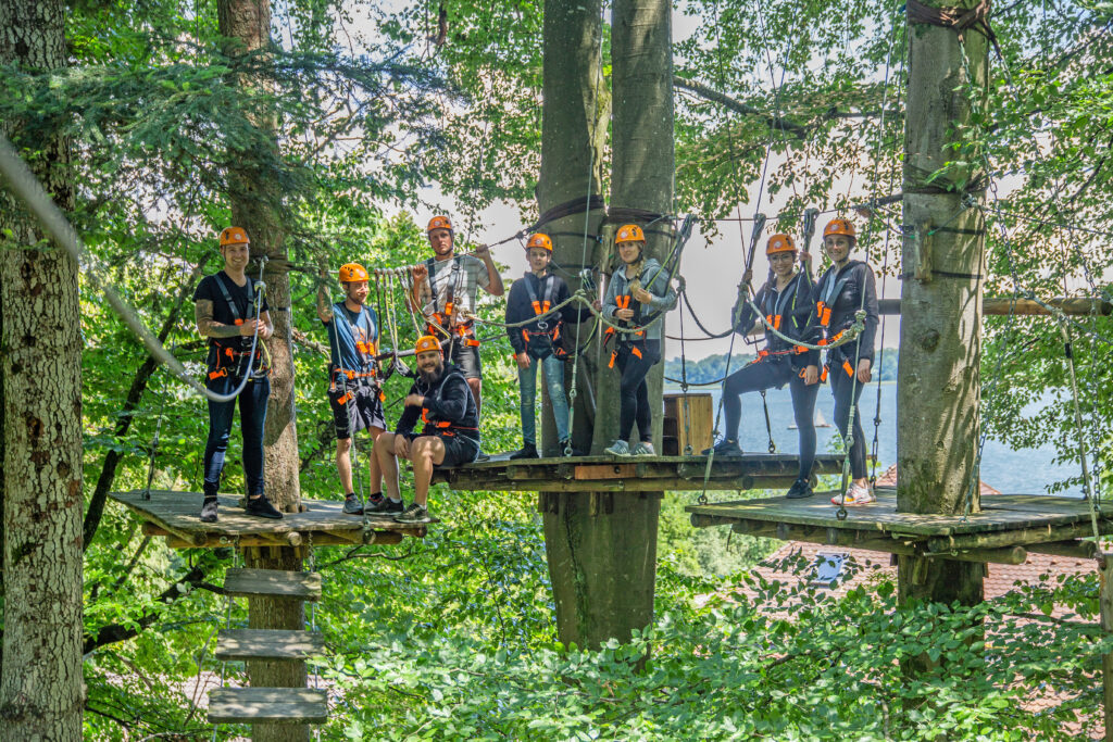 Eine große Gruppe an Personen ist im Kletterwald und klettert. Im Hintergrund ist der Chiemsee zwischen den Bäumen zu sehen.