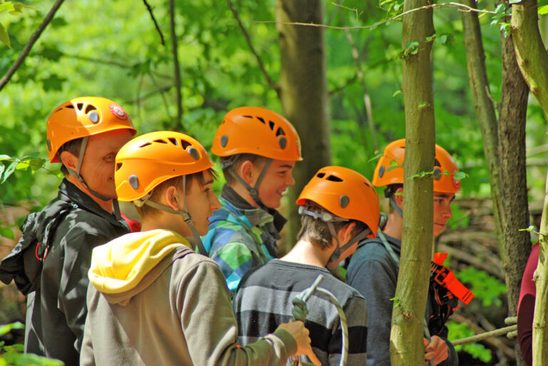 Eine Gruppe Kinder machen sich bereit zum Klettern im Kletterwald. Sie tragen bereits Kletterausrüstung und einen orangenen Kletterhelm. Im Hintergrund steht der Lehrer.