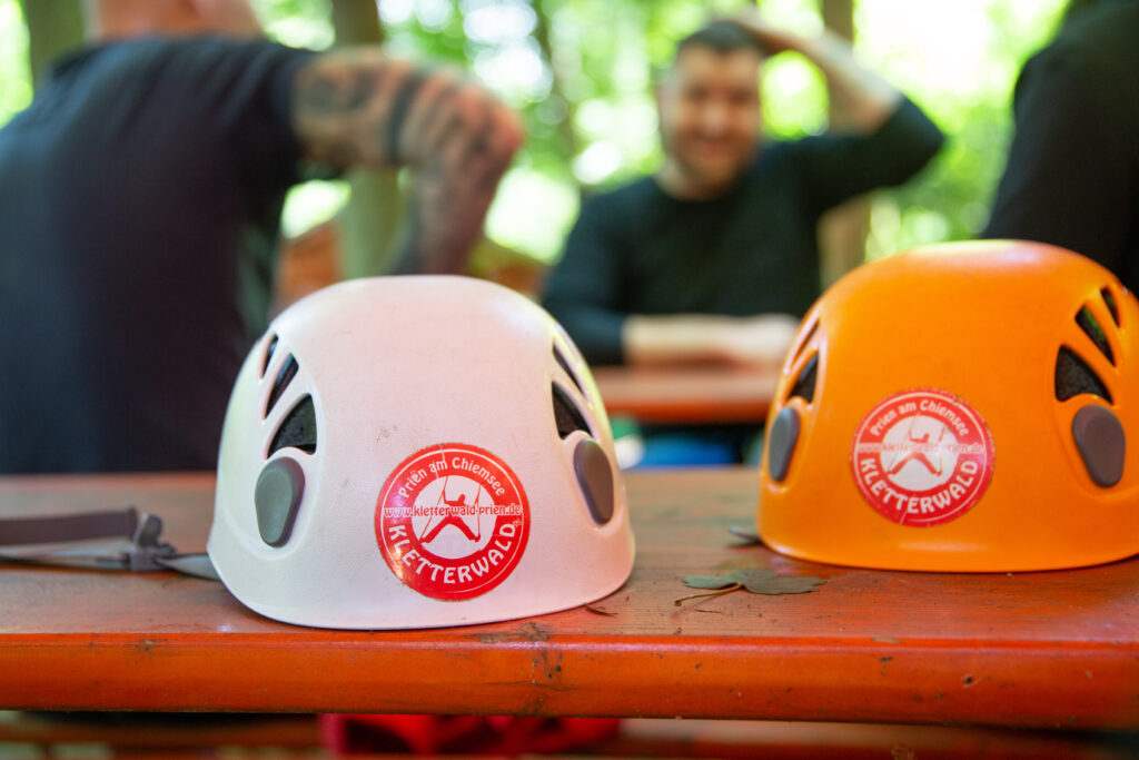 Zwei Kletterhelme liegen nebeneinander auf einer Bierbank. Ein Helm ist weiß, der andere orange. Auf beiden ist ein Kletterwald Logo abgebildet.
