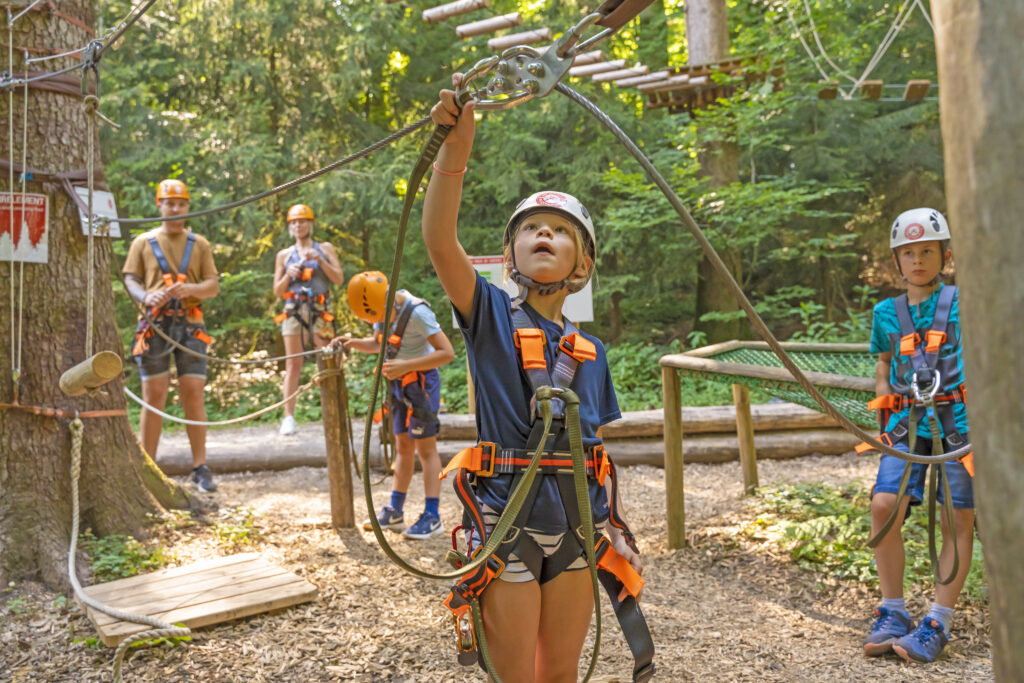 Ein junges Mädchen klettert im Kletterwald und gefästigt gerade ihren Karabiner in dem Kletterdraht. Sie schaut konzentriert. Im Hintergrund sind weitere Kletterer zu sehen.