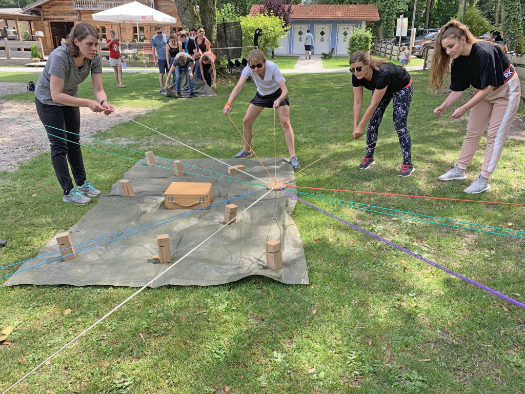 Gruppe von Menschen steht auf einer Wiese im Kreis und arbeitet gemeinsam mit gespannten Seilen an einer Aufgabe in der Mitte.