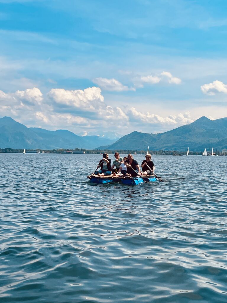 Fünf Personen sitzen auf einem selbstgebauten Floß und fahren damit an einem sonnigen Tag auf dem Chiemsee. Im Hintergrund sind die Alpen.