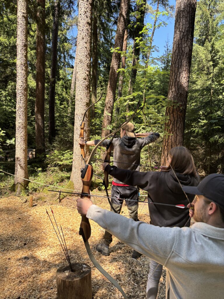 3 Personen haben einen Bogen in der Hand sowie einen Pfeil, um Bogenzuschießen. Sie stehen im Münchner Wald Kletterwald Vaterstetten.