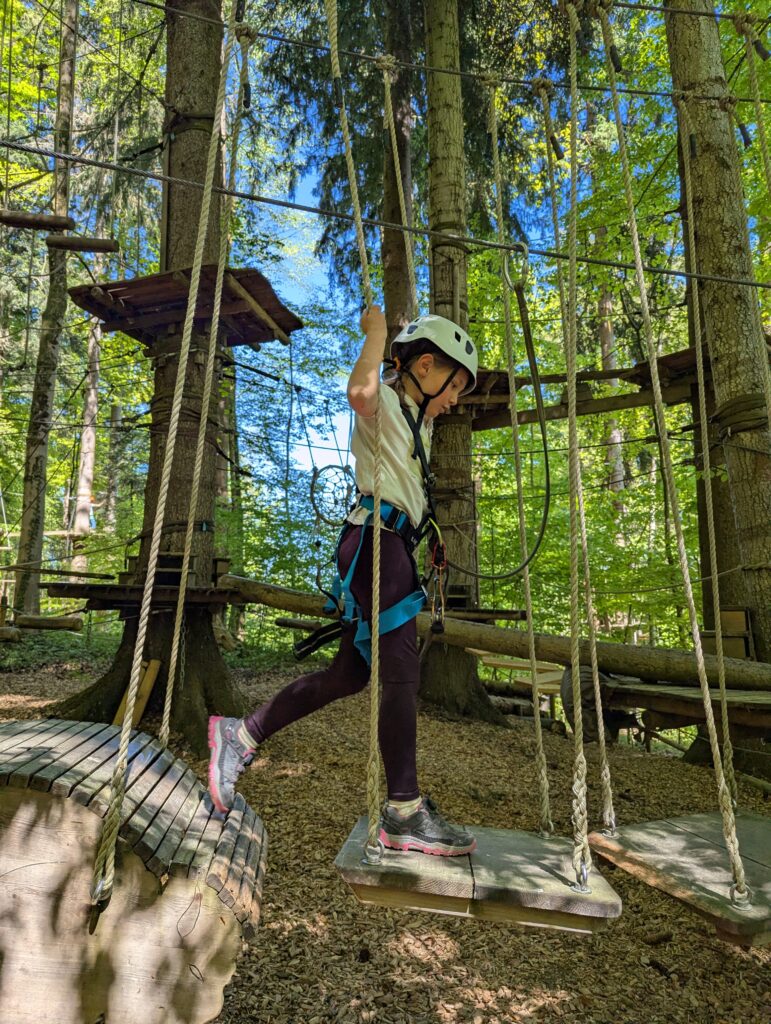 Ein Mädchen mit weißem Helm klettert in einem Parcours im Kletterwald Prien am Chiemsee. Der Parcour enthält an Seilen hängende Plattformen.