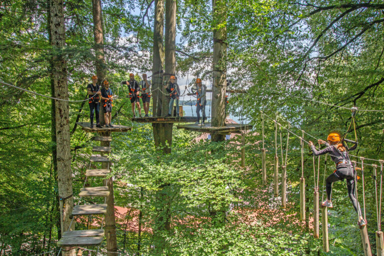 Sieben Personen klettern einen Parcours im Kletterwald und haben viel Spaß. Gut zu erkennen ist der Kletterwald Prien am Chiemsee und auch der Chiemsee ist im Hintergrund zu erkennen.