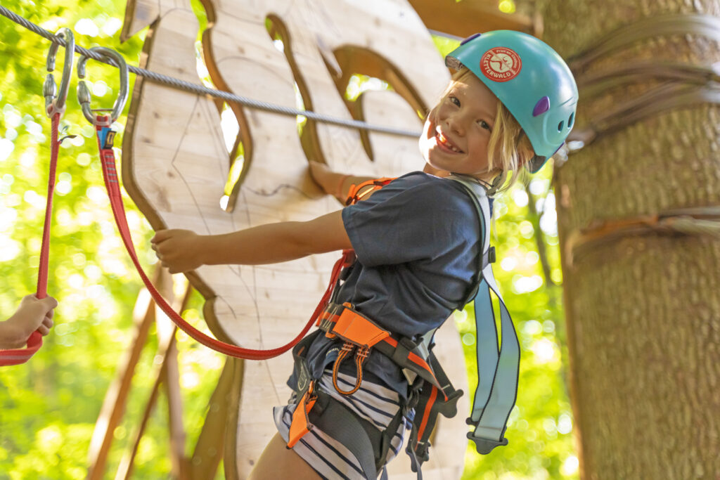 Ein junges Mädchen mit einem blauen Helm klettert einen Kinderparcours im Kletterwald iin Prien am Chiemsee und lächelt direkt in die Kamera