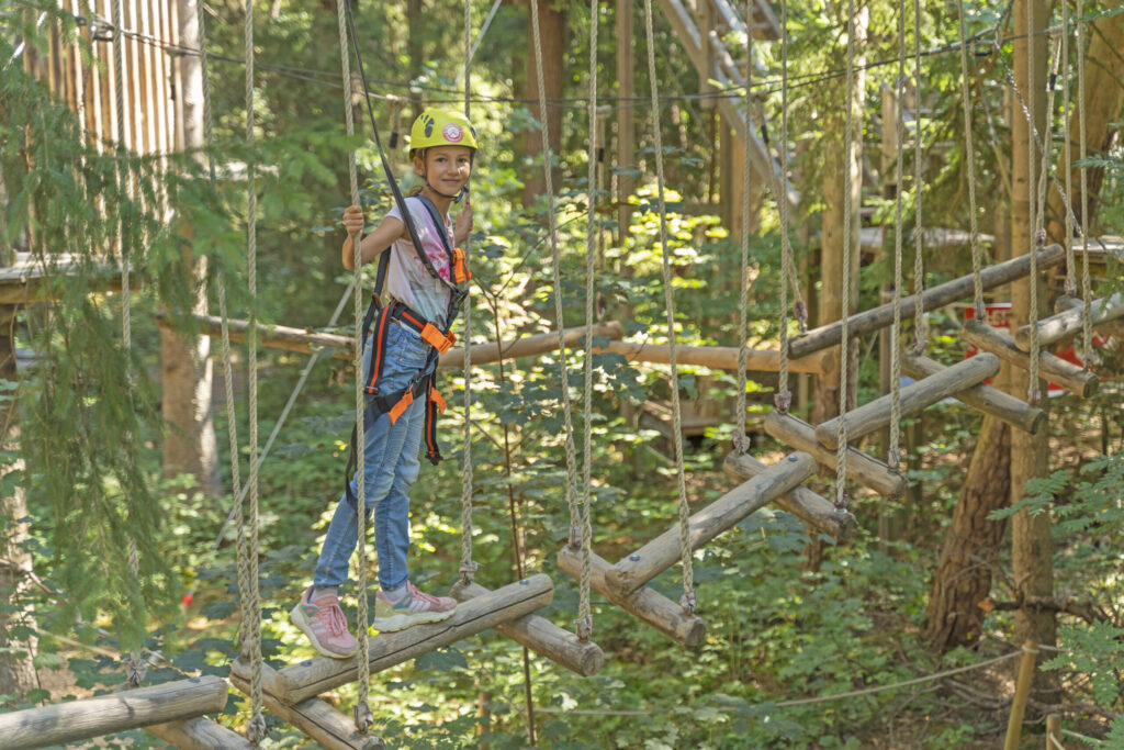 Ein Mädchen steht auf hängenden Baumstämmen in einem Kletterwald Parccours im Münchner Wald. Im Hintergrund ist der Kletterwald Prien am Chiemsee.