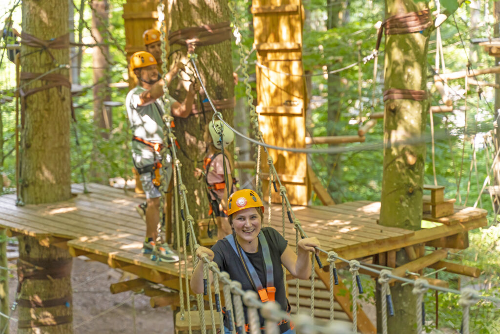 Eine Frau klettert durch einen Parours, im Hintergrund hilft ein Vater seiner Tochter mit der Sicherung.