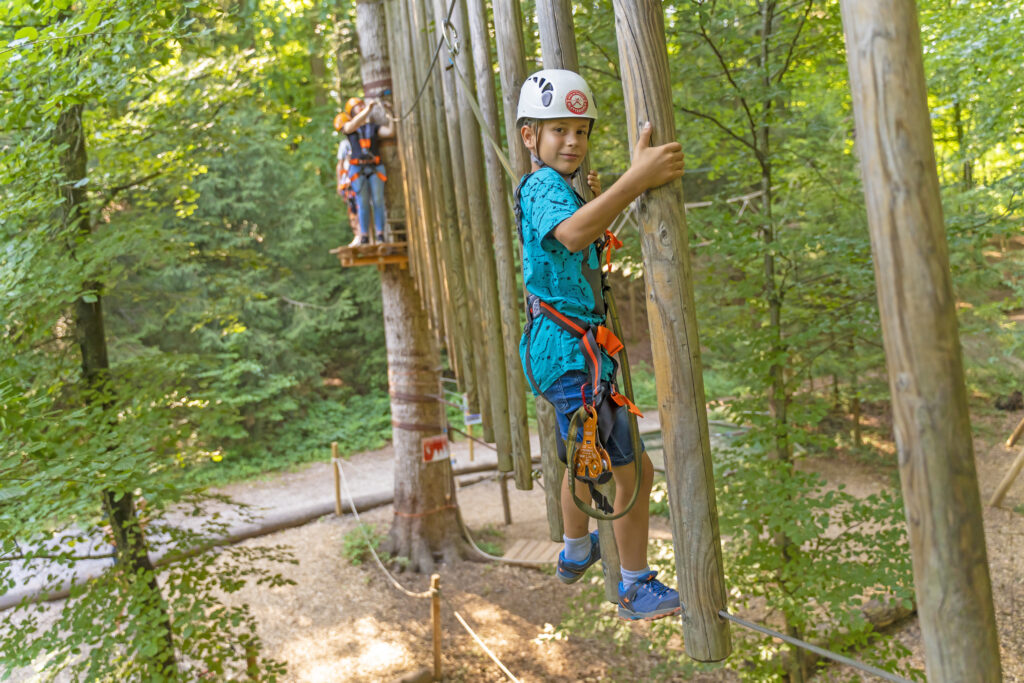 Ein Junge klettert einen Parcours im Kletterwald Prien und lacht diekt in die Kamera. Er hält sich an Baumstämmen fest und im Hintergrund warten weitere Gäste.