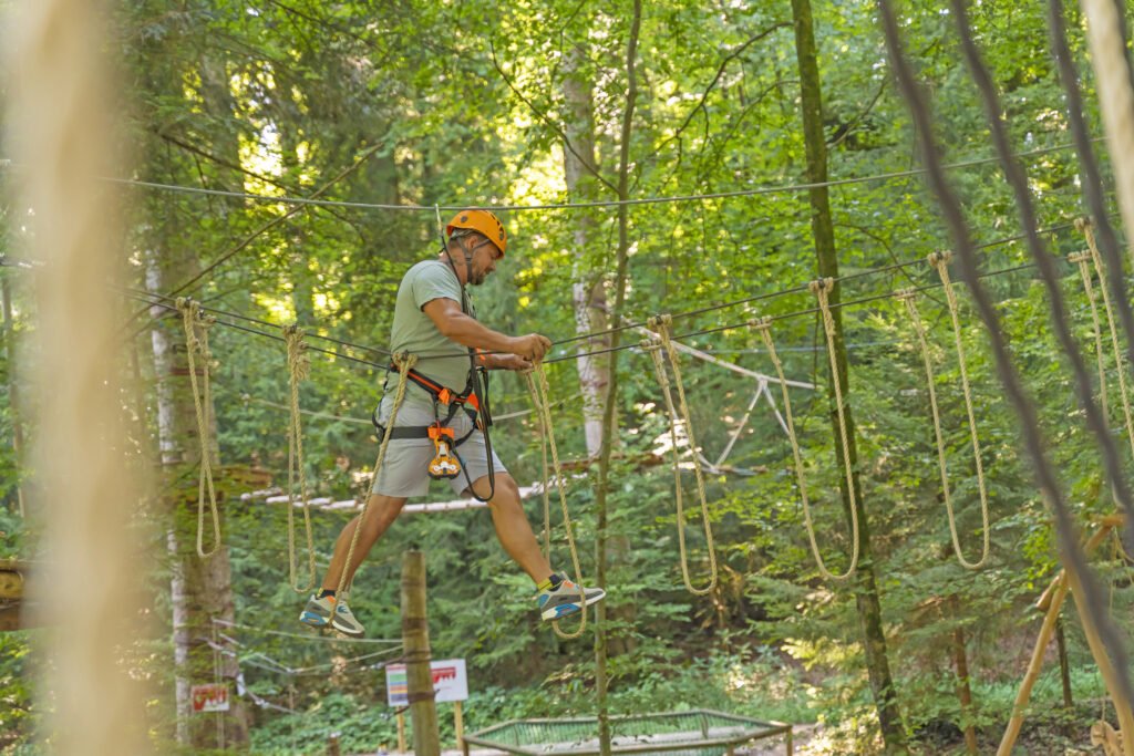Ein Mann überquert eine Übung im Kletterwaldparcours, welcher aus Seilen besteht.Im Hintergrund sieht man weitere Parcours im Ketterwald Prien am Chiemsee.