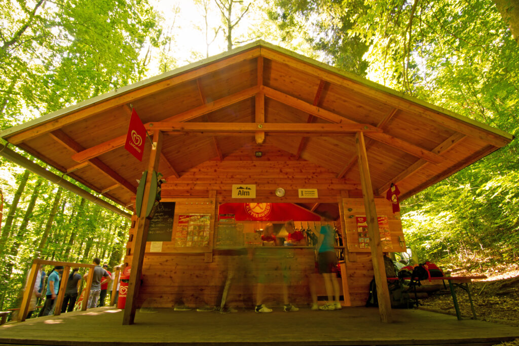 Eine Hütte aus Holz mit mit einem Kiosk hinter dem Fenster. Im Hintergrund ist grüner Wald zu sehen.